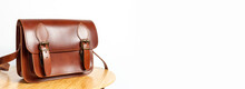 Fashionable Brown Women's Bag Made Of Genuine Leather On Wooden Table Against White Wall With Place For Text. Fashion Concept. Details Of Leather Bag Belt Metal Buckle Clasp. Stylish Female Accessory