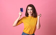 © NaMong Productions - Young woman holding credit card  in casual yellow shrit with gorgeous in trust and confidence for money transaction isolated on bright pink background.