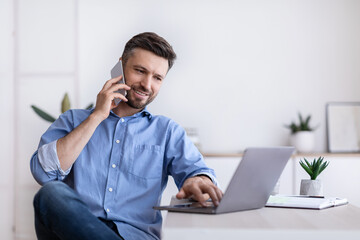 Wall Mural - Cheerful Young Man Working Remotely From Home Office With Cellphone And Laptop