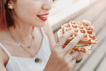 Wall Mural - Smiling hipster woman eating a delicious sandwich with a loaf of a checkered Viennese waffle. Modern Street food concept