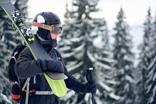 Portrait Photo Of Male Skier Standing On Slope Top, Looking At Distance In His Goggles. Professional Sportsman In Helmet With Pair Of Skis. Grey Snowy Pine Trees On Blurred Background. Monochrome View