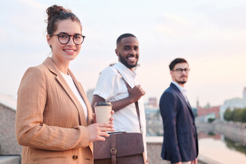 Portrait of positive adult students standing in row, focus on attractive girl in glasses drinking coffee outdoors