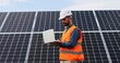 Portrait of electrician in safety helmet and uniform using laptop checking solar panels. Male technician at solar station.