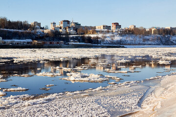 Ice drift on a river with blue high water and big water, white snow broken ice full of hummocks in it and city with houses at a background in sunny spring day.