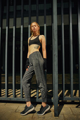 Wall Mural - Young fit woman in sport suit stands by metal fence oudoors