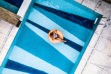 Aerial View Of Tanned Woman Floating Comfortably On Orange Inflatable Blow Up Ring In Luxury Villa Swimming Pool