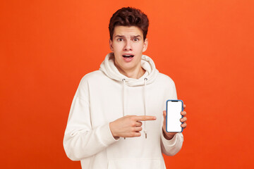 Wow! Shocked excited teenager in casual white hoodie showing smartphone with empty blank, new favorable tariffs for calls. Indoor studio shot isolated on orange background