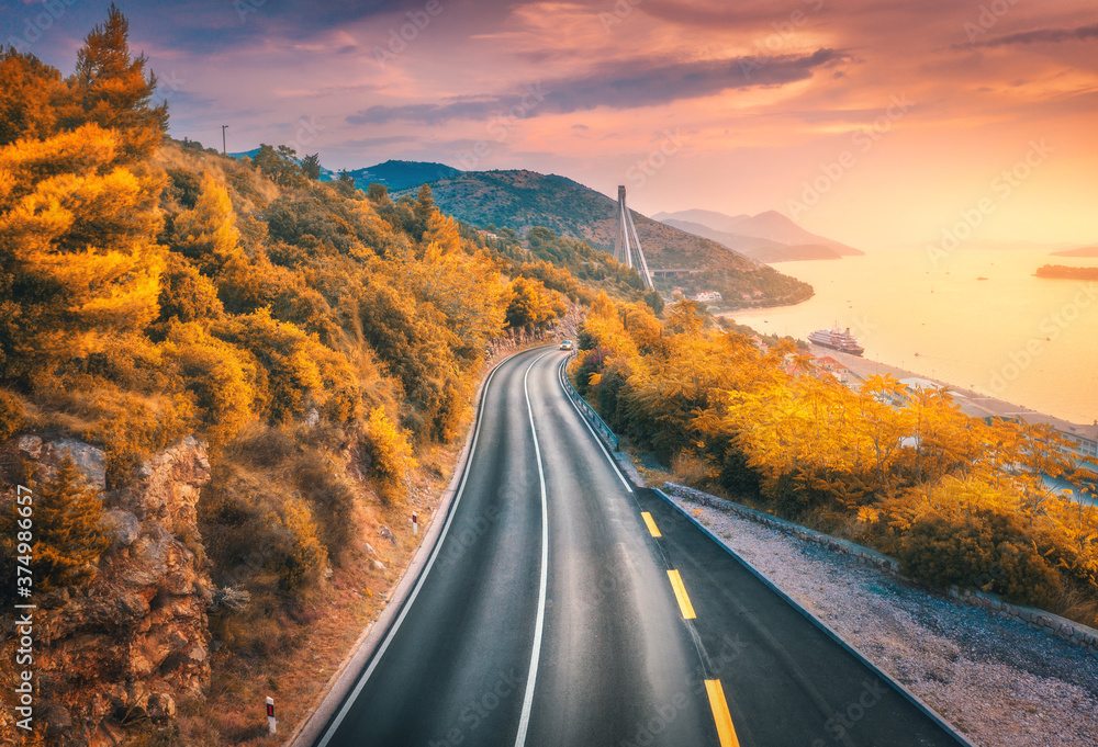 Aerial view of mountain road and beautiful orange forest at colorful sunset in autumn. Dubrovnik, Croatia. Top view of road, trees, sea, mountain, red sky in fall. Landscape with highway and sea coast - obrazy, fototapety, plakaty 