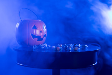 A plastic pumpkin jack o lantern bucket for trick or treating with saltwater taffy on a table against a back background with fog