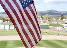 The American Flag Waving With A Rural Town's Baseball Game In The Background.