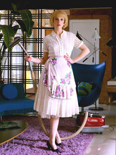 Woman Dressed In Vintage Attire Holding A Vacuum In One Hand And Drink In The Other In A Mid-century Modern Home.