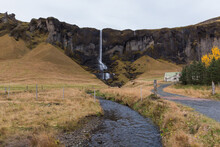 Road To The Mountains In Iceland. Tall Waterfall And River Cuts Through A Farmland 