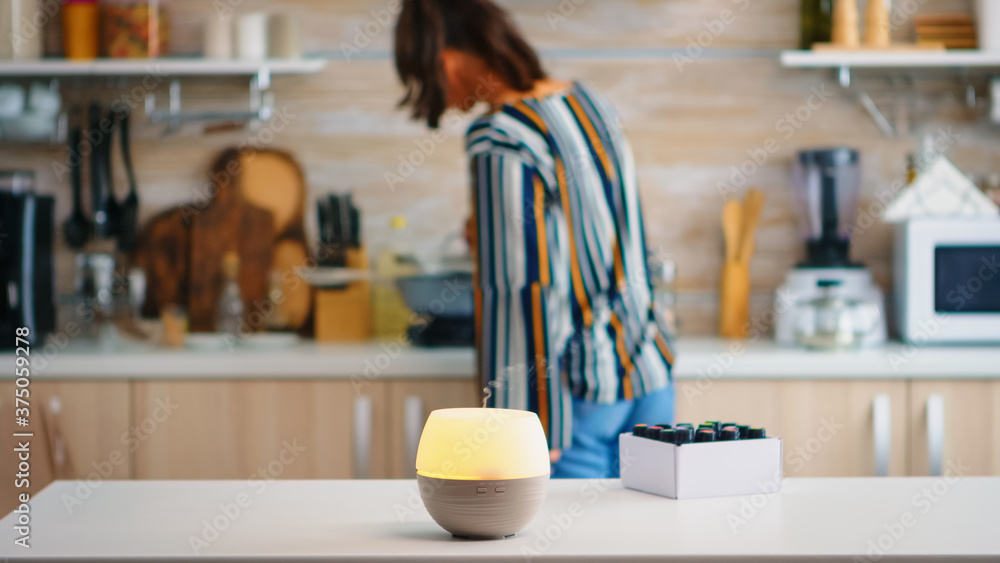 Essential oils diffuser distributing aromatherapy while woman walks in the kitchen. Aroma health essence, welness aromatherapy home spa fragrance tranquil theraphy, therapeutic steam, mental health - obrazy, fototapety, plakaty 