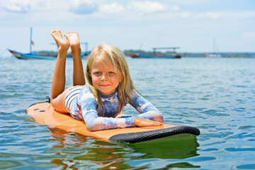 Happy baby girl - young surfer learn to ride on surfboard with fun on sea waves. Active family lifestyle, kids outdoor water sport lessons, swimming activity in surf camp. Summer vacation with child.