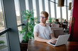 © александр таланцев - Young guy in a white t-shirt and glasses works on a laptop sitting at a table in a cafeteria
