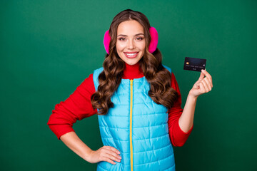 Poster - Photo of pretty lovely young lady smiling showing credit card hand hip bank promotion profitable deposit saving wear pink ear warmers blue vest red sweater isolated green color background