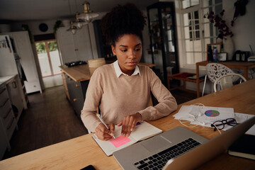 Wall Mural - Young freelancer writing details on book while working on laptop at home during lockdown