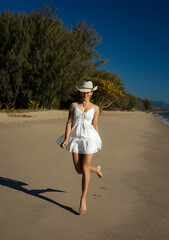 Wall Mural - A beautiful young curly-hair woman in a little white/beige dress and cowboy's straw hat  is posing at the beach. Portrait Photography.