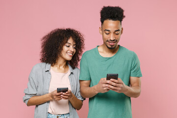 Wall Mural - Smiling young african american couple two friends guy girl in gray green casual clothes posing using mobile cell phone typing sms message isolated on pastel pink color wall background studio portrait.