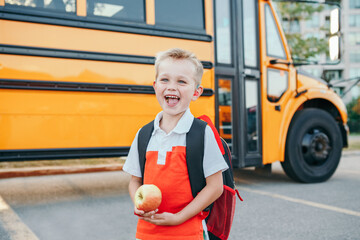 Caucasian boy student with backpack near yellow bus on first September day. Child kid eating apple fruit at school yard outdoors. Education and back to school in Autumn Fall.