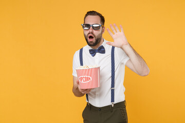 Wall Mural - Shocked scared young bearded man 20s wearing white shirt bow-tie suspender 3d glasses watching movie film, holding bucket of popcorn covering with hand isolated on yellow background studio portrait.