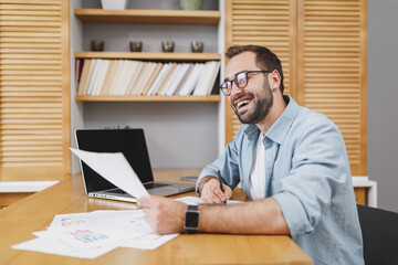 Wall Mural - Cheerful smiling funny young bearded business man 20s wearing blue shirt glasses sitting at desk with papers document working on laptop pc computer writing in notebook at home or office.