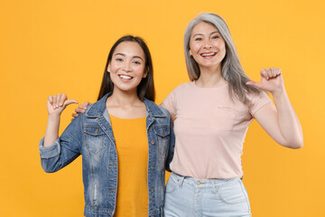 Wall Mural - Smiling cheerful family asian women girls gray-haired mother and brunette daughter in casual clothes posing hugging pointing thumbs on themselves isolated on yellow color background studio portrait.