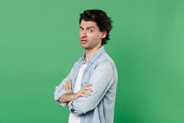 Side view of puzzled perplexed young brunet man 20s wearing casual clothes white t-shirt denim shirt posing standing holding hands crossed looking camera isolated on green background studio portrait.