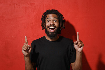 Wall Mural - Amazed young african american man with dreadlocks 20s in black casual t-shirt posing pointing index fingers up on mock up copy space isolated on bright red color wall background studio portrait.