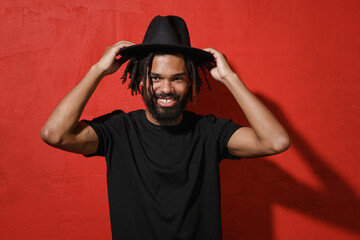 Wall Mural - Smiling handsome attractive young african american man guy with dreadlocks 20s in black casual t-shirt posing hold hat on head looking camera isolated on bright red color background studio portrait.