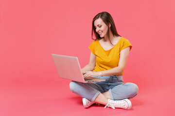 Full length portrait of beautiful smiling attractive young brunette woman 20s wearing yellow casual t-shirt sitting on floor working on laptop pc computer isolated on pink color background studio.