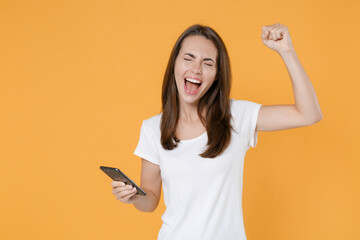 Happy young brunette woman 20s in white blank empty design casual t-shirt using mobile cell typing sms message doing winner gesture keeping eyes closed isolated on yellow background studio portrait.