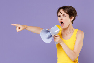 Angry young brunette woman girl in yellow casual tank top posing isolated on pastel violet wall background studio portrait. People lifestyle concept. Screaming in megaphone point index finger aside.