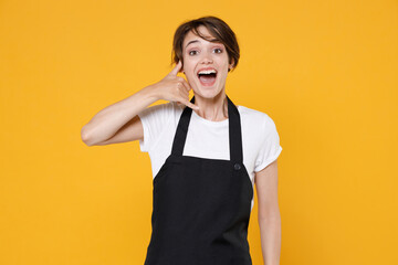 Excited young female woman 20s barista bartender barman employee in white t-shirt apron working in coffee shop doing phone gesture like says call me back isolated on yellow background studio portrait.