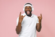 © ViDi Studio - Excited young bearded african american fitness sports man in headband t-shirt listen music with headphones depicting heavy metal rock sign horns up isolated on pink color background studio portrait.
