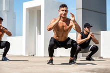 Group Of Fit Sports Men Doing Squat Bodyweight Workout Training Outdoors On Building Rooftop - Exercise In The Open Air Concept