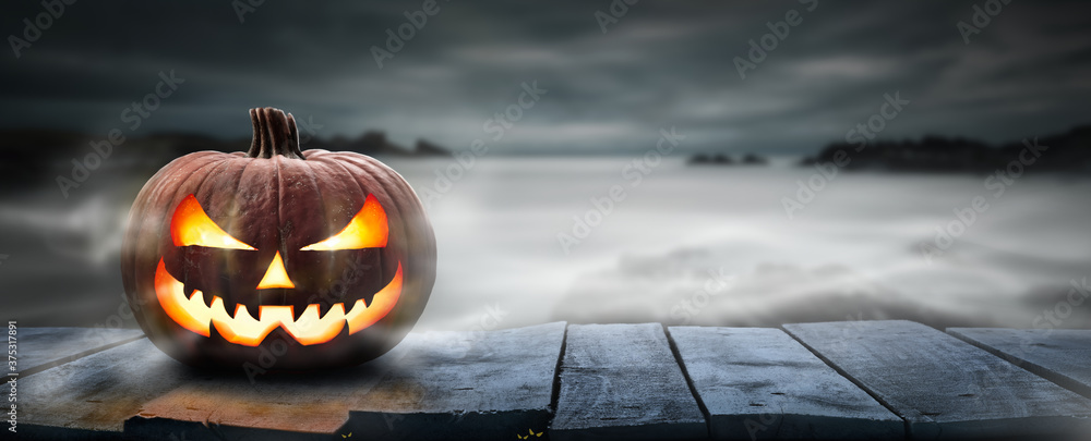 One spooky halloween pumpkin, Jack O Lantern, with an evil face and eyes on a wooden bench, table with a misty gray coastal night background with space for product placement. - obrazy, fototapety, plakaty 