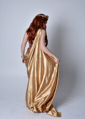 Wall Mural - Full length portrait of girl with red hair wearing long grecian toga and golden wreath. Standing pose with back to the camera,  isolated against a grey studio background.