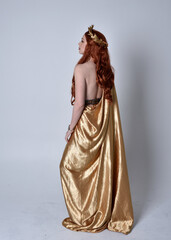 Wall Mural - Full length portrait of girl with red hair wearing long grecian toga and golden wreath. Standing pose with back to the camera,  isolated against a grey studio background.