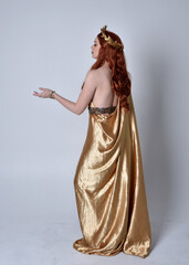 Wall Mural - Full length portrait of girl with red hair wearing long grecian toga and golden wreath. Standing pose with back to the camera,  isolated against a grey studio background.