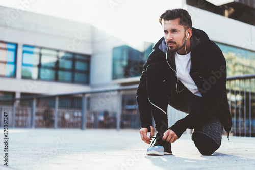 Determined male jogger in tracking suit earphones checking new cross sneakers for jogging prepare for training outdoors, serious caucasian sportsman listening music and tying laces on shoes