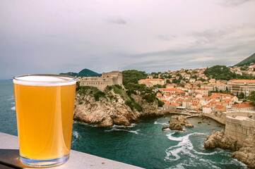 Wall Mural - Glass of light beer against view of blue sea and rocky coast background. Dubrovnik old town surrounded by old walls. View from above of red rooftops, roofs and fortress.