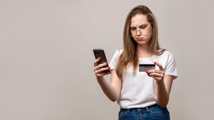 Bank fraud. Mobile scam. Confused woman in glasses using phone credit card isolated on gray copy space background. Cashless payment. Personal data verification.