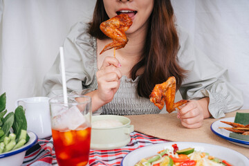 Asian woman eating roast chicken stick with both hands with thai tea.