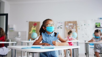 Canvas Print - Small girl with face mask sitting on lesson indoors in classroom, writing.