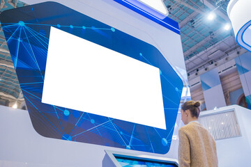 Woman using electronic kiosk and looking at white blank large interactive wall display in dark room of modern technology exhibition. Mock up, futuristic, template, education and technology concept