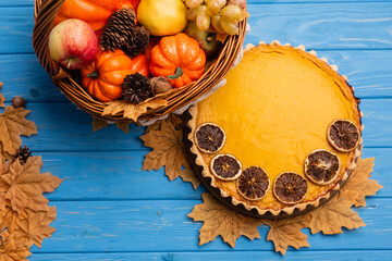 Wall Mural - top view of autumnal harvest in wicker basket near pumpkin pie on blue wooden background