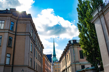 Wall Mural - Sun Peeking over a Building Lining an Alley Way with the Steeple of the German Church on the Horizon in the Gamla Stan Neighborhood of Stockholm, Sweden