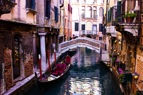 Beautiful Venice canal with gondola and red brick bridge. The luxurious gondola has red velvet-lined seats. On one wall there are lit lights and purple flowers in the balconies. © Julia Lopatina