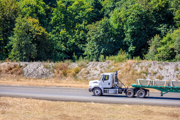 Big rig day cab semi truck transporting fixed with slings commercial cargo on flat bed semi trailer running on the local road with forest on the side